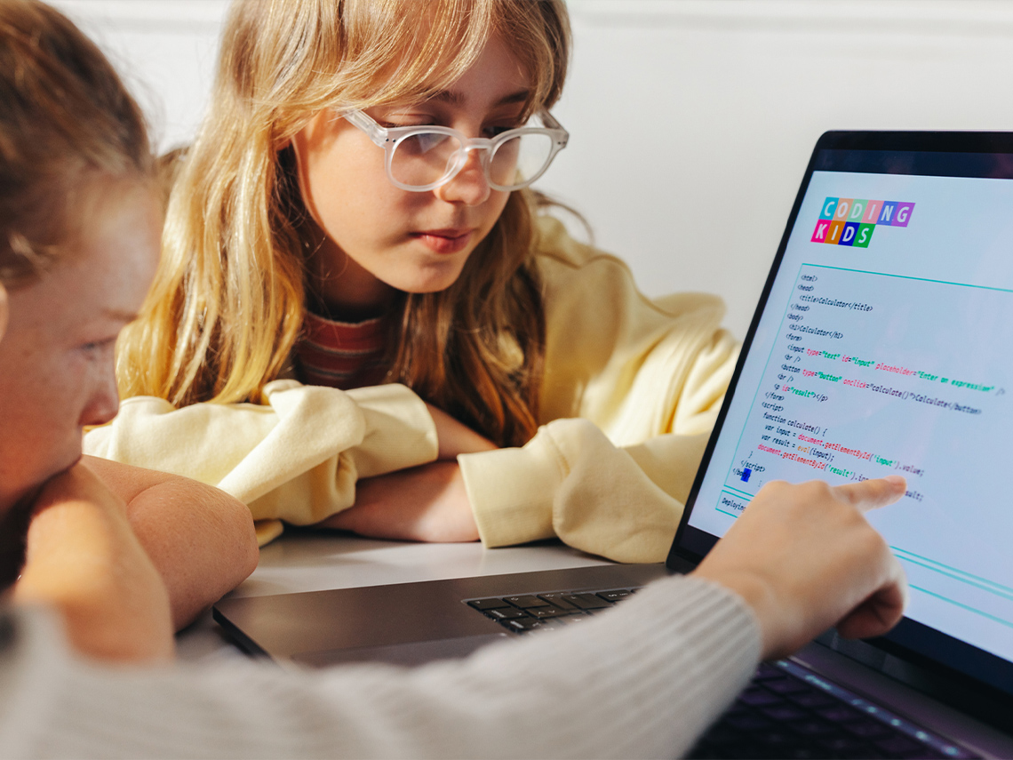 Two students sitting in front of a laptop with code displayed on the screen. One of the students is pointing to a line of code.