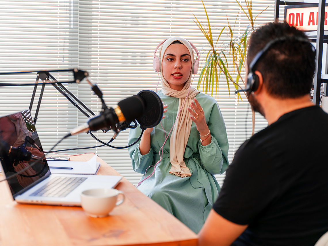 There are two adults recording a podcast. They are wearing headphones and there is a large microphone between them.