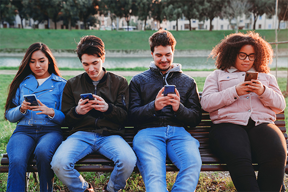 A group of teenagers sitting on bench all on their phones.
