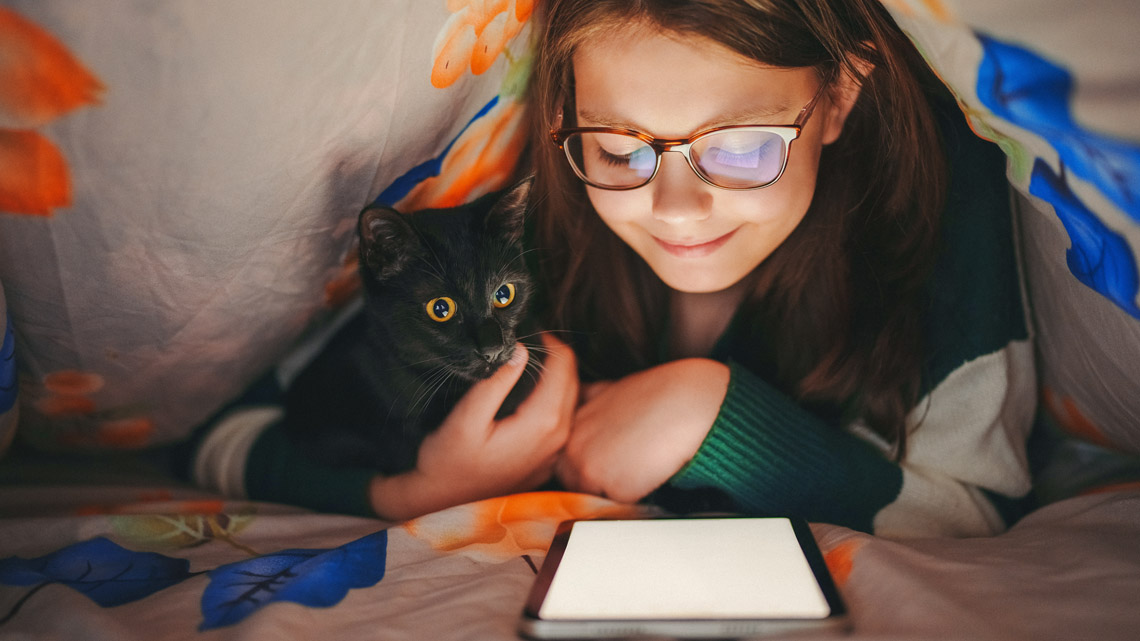 A girl reading on a tablet under the covers.