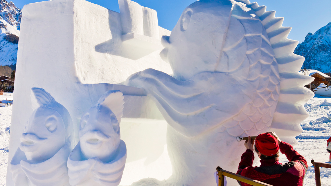 A person using tools to sculpt snow into a decorative fish sculpture.