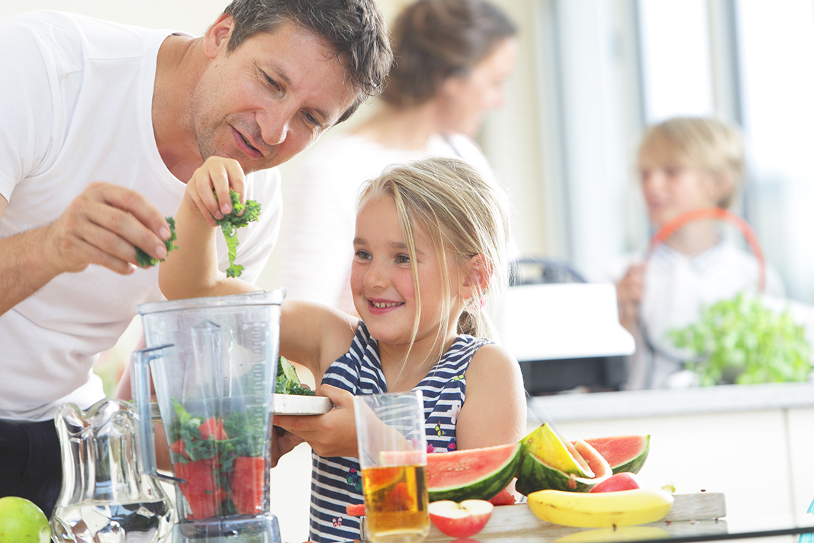 A father and daughter putting fruit and vegetables in a blender jar.