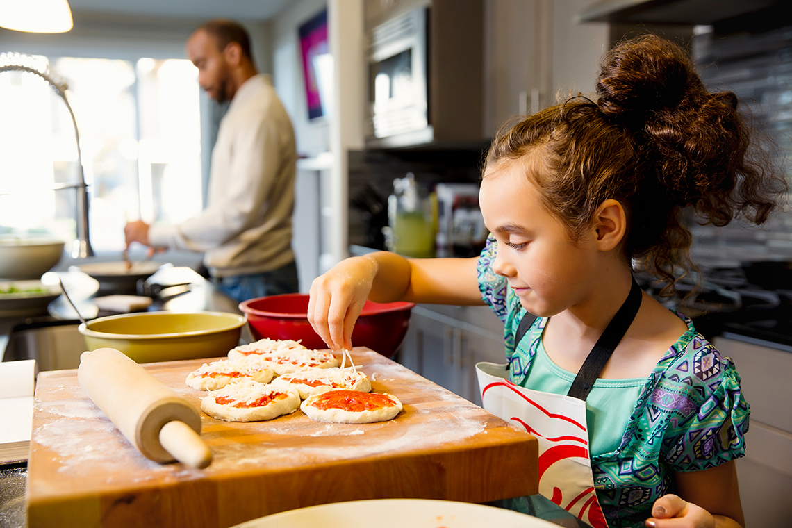 A girl in a kitchen putting shredded cheese on mini pizzas.