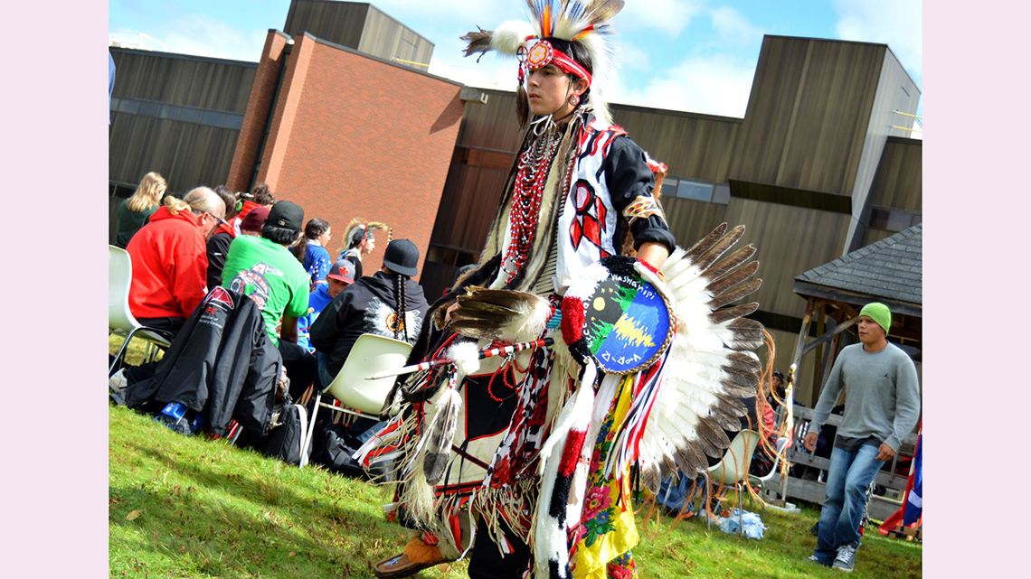 Someone performing a powwow dance with an audience in the background.