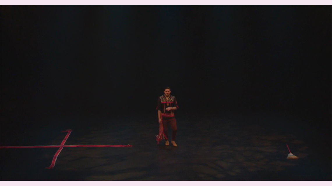 A Métis individual stands positioned to dance on a darkened stage with ribbons on one side and a broom on the other side.