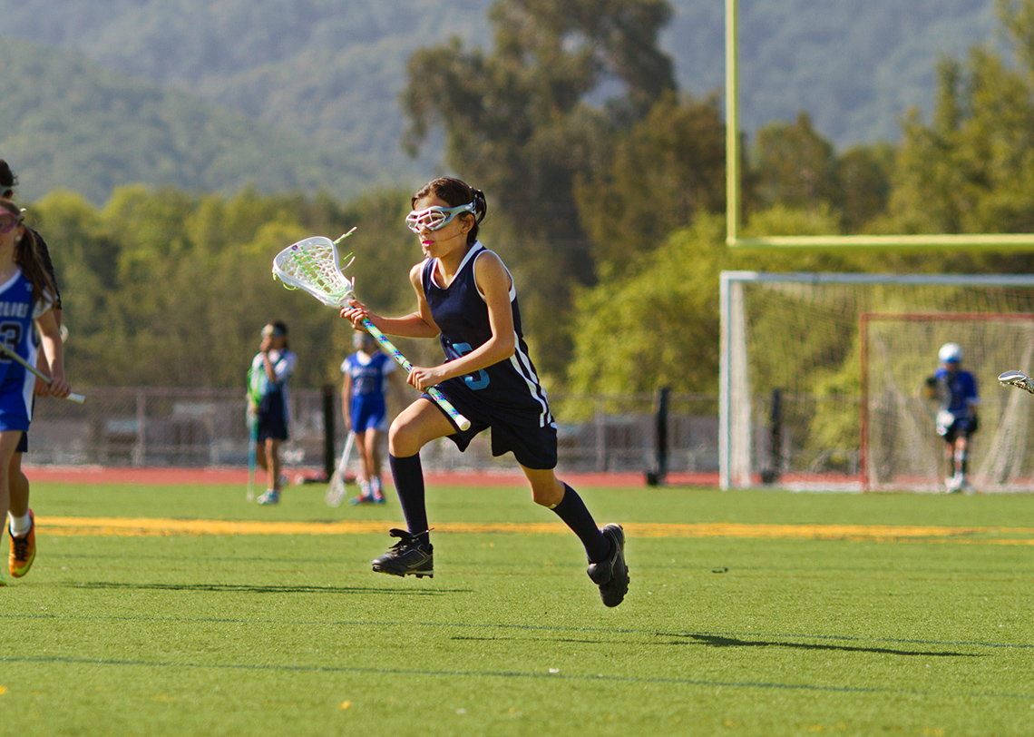 A girl wearing cleats and a uniform stands on a field carrying a lacrosse stick and wearing goggles while other players and a net are in the background.