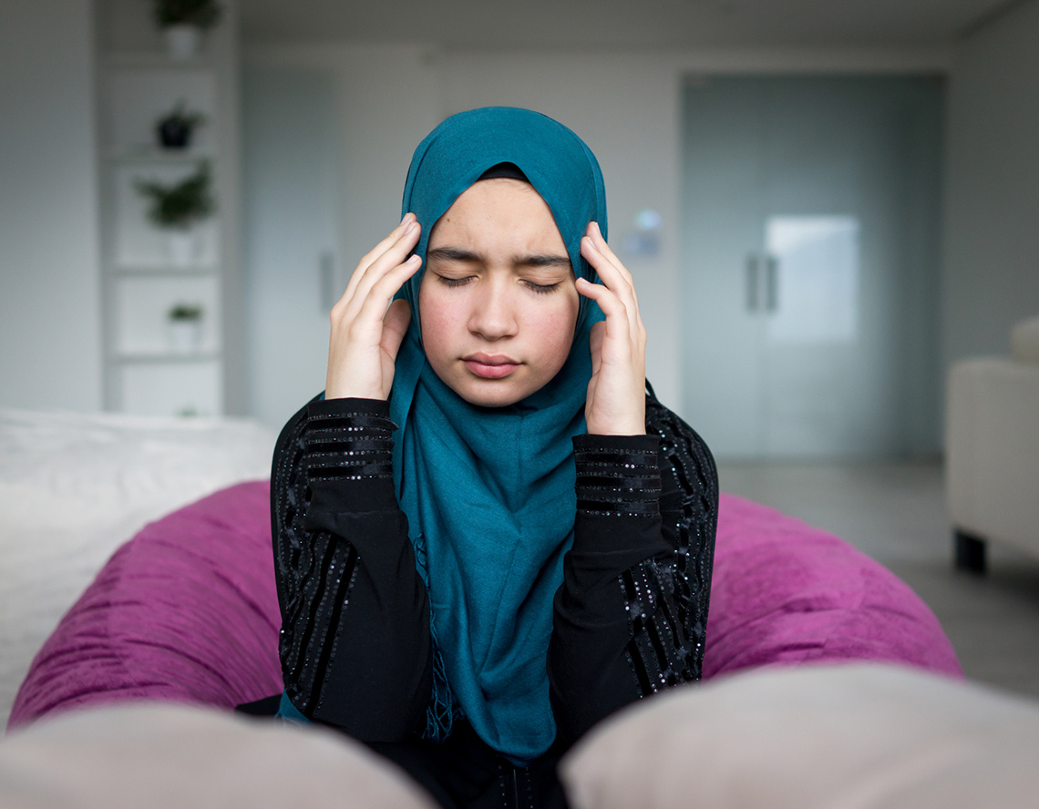 A girl sitting on cushions with her eyes closed and her fingers on her temples.