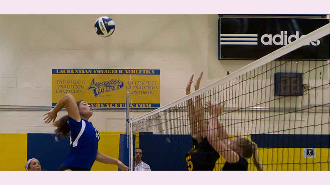 Action shot of a volleyball game in a school gym where one of the female players is about to spike the ball while the opposing team has their hands raised to block.