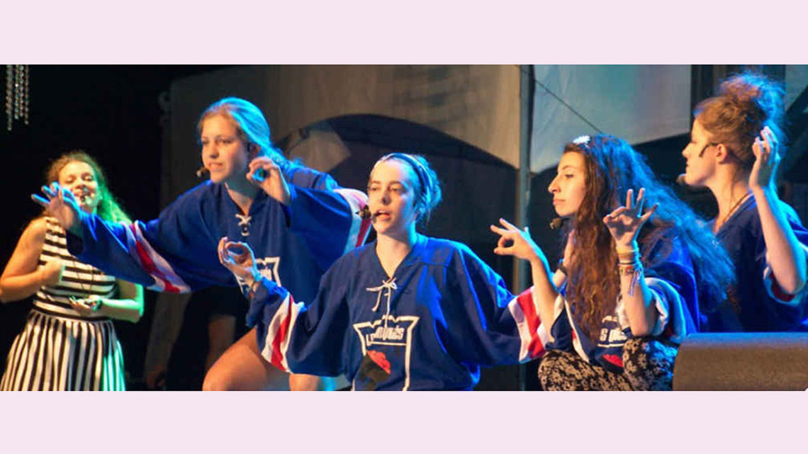 Five teen actors perform a play onstage wearing microphone headsets. Three of the actors are wearing matching hockey-style jerseys and are making similar hand signs where their index fingers and thumbs are connected to form the shape of a circle.