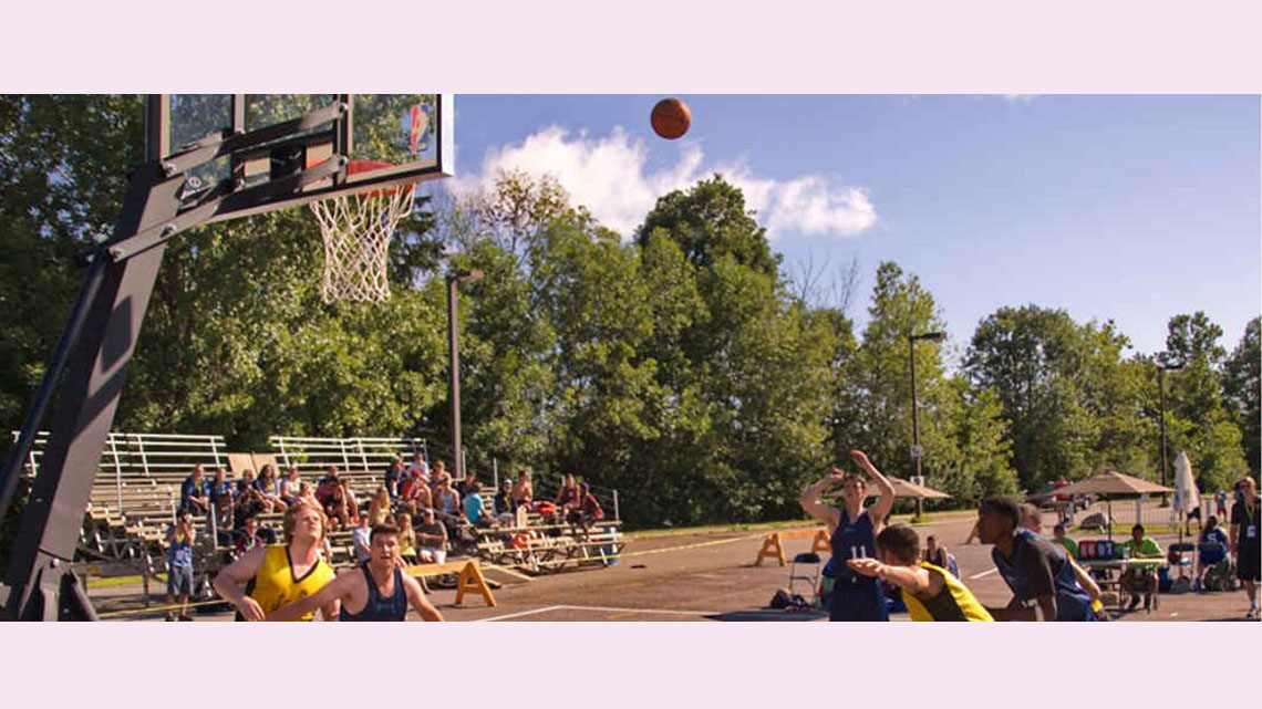 An outdoor basketball court with a game in progress. One of the male players is mid-shot while several others play offence and defence.