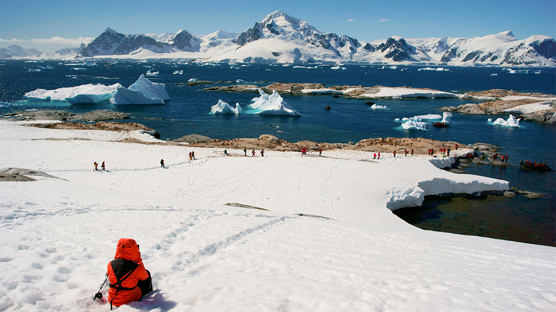 A polar region with snow on the ground, icebergs in the water and snowy mountains.