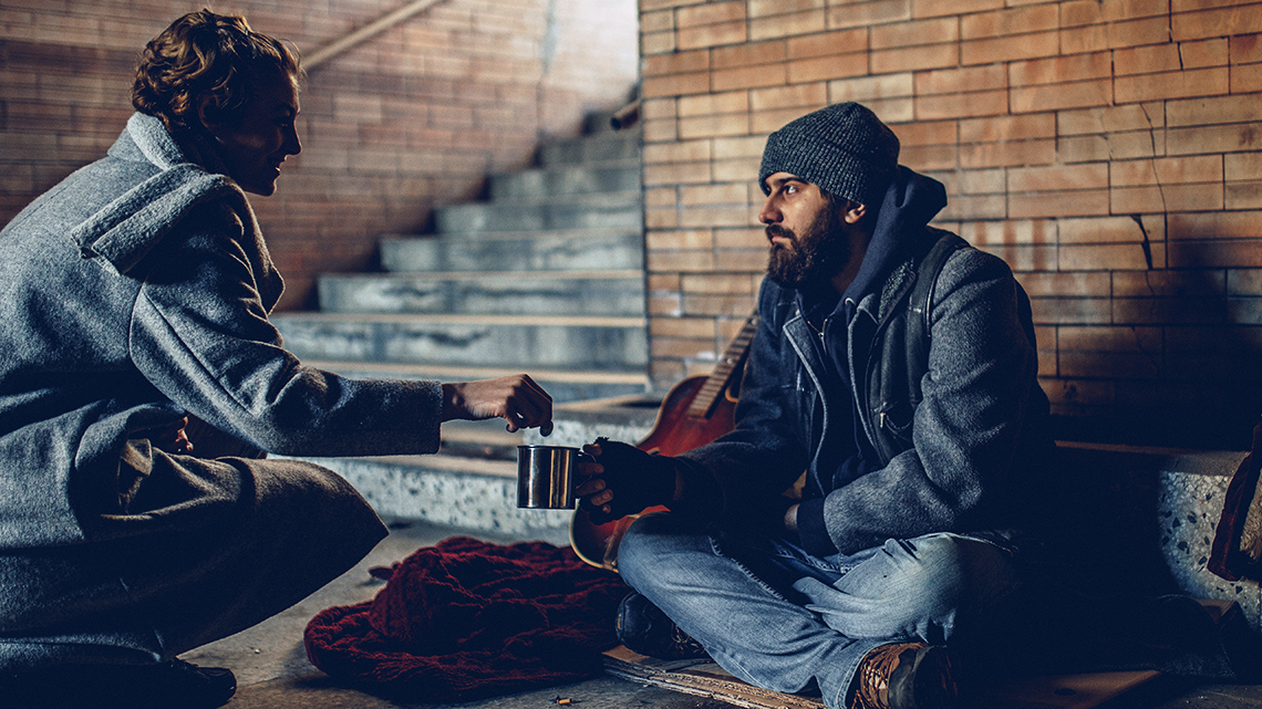 A woman puts change in a cup held by a young man sitting on cardboard on the ground.