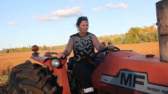 A woman on a tractor is working outside on a hot, dry day with brown crops behind her.