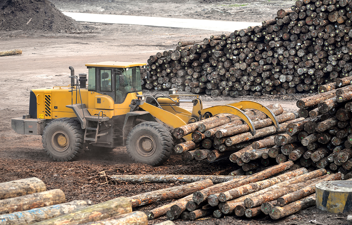 A construction vehicle is stacking cut down trees on a flat plain of dirt.