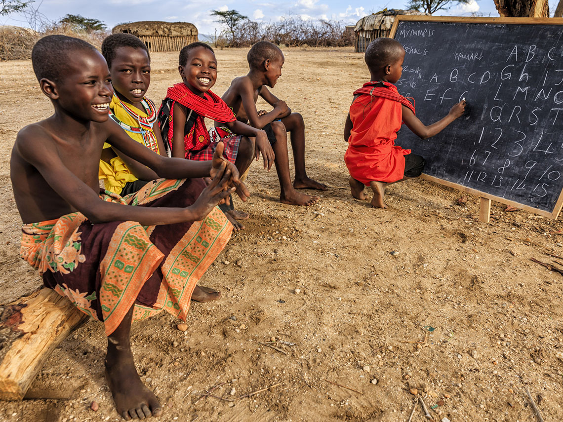 Children on a log in Kenya, Africa watch another child write the English alphabet on a chalkboard.