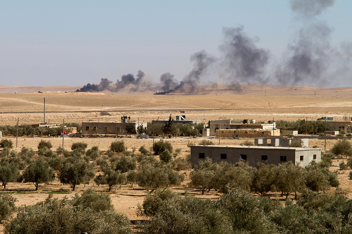 A desert with trees and buildings in Syria. Background has black smoke coming from a battle.
