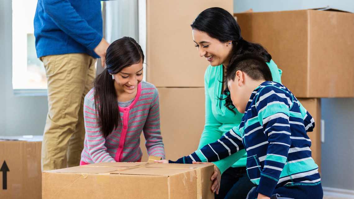 A family moving into a house A room full of unopen moving boxes. A person pushes their child in an empty box.