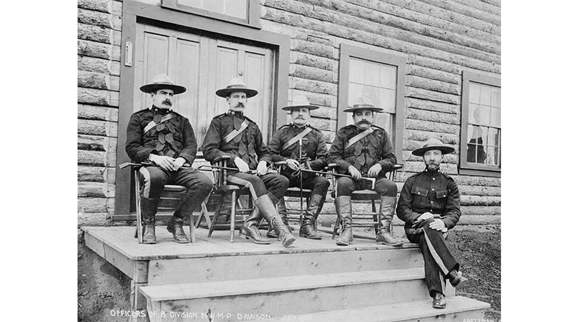 North-West Mounted Police dressed in uniform with a sash, a necktie, wide brim hats, and boots. Each person is male and of European descent.