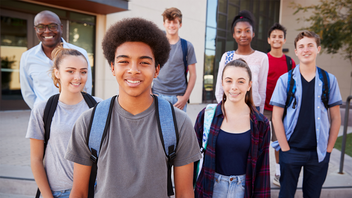 A multi-ethnic group of students smiling. Their teacher is standing behind them smiling as well.