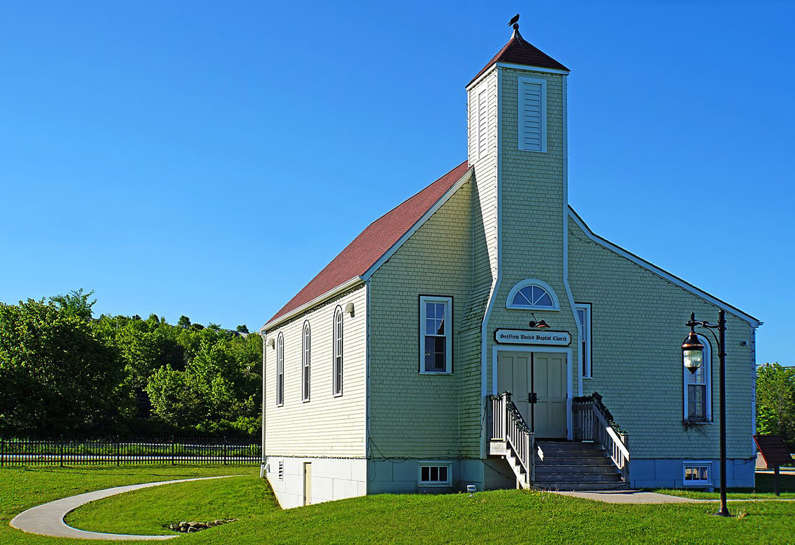A small church with a steep pitched roof and a steeple. It sits in a grassy area surrounded by open sky and trees