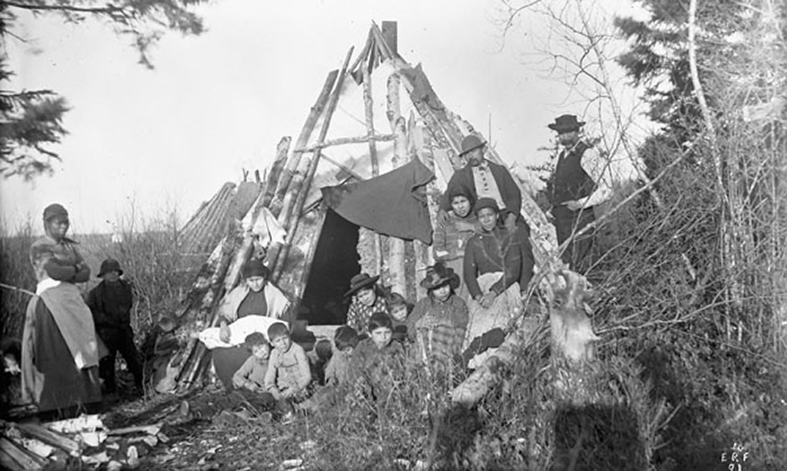 A group of Indigenous individuals standing in front of a tipi. There are trees around them.