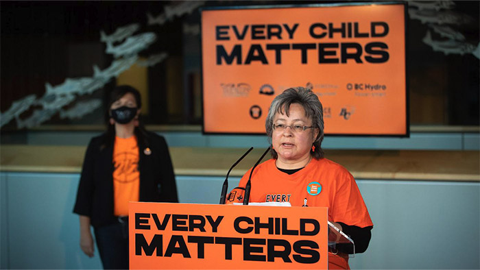 A woman in orange shirt speaks at a podium surrounded by banners reading “Every Child Matters”