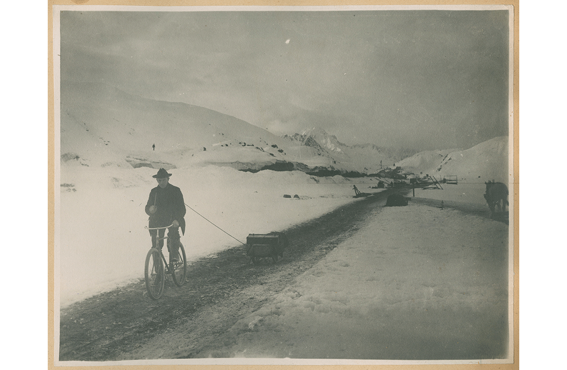 A man on a bicycle pulling sled down a road in between two high, snowy hills.