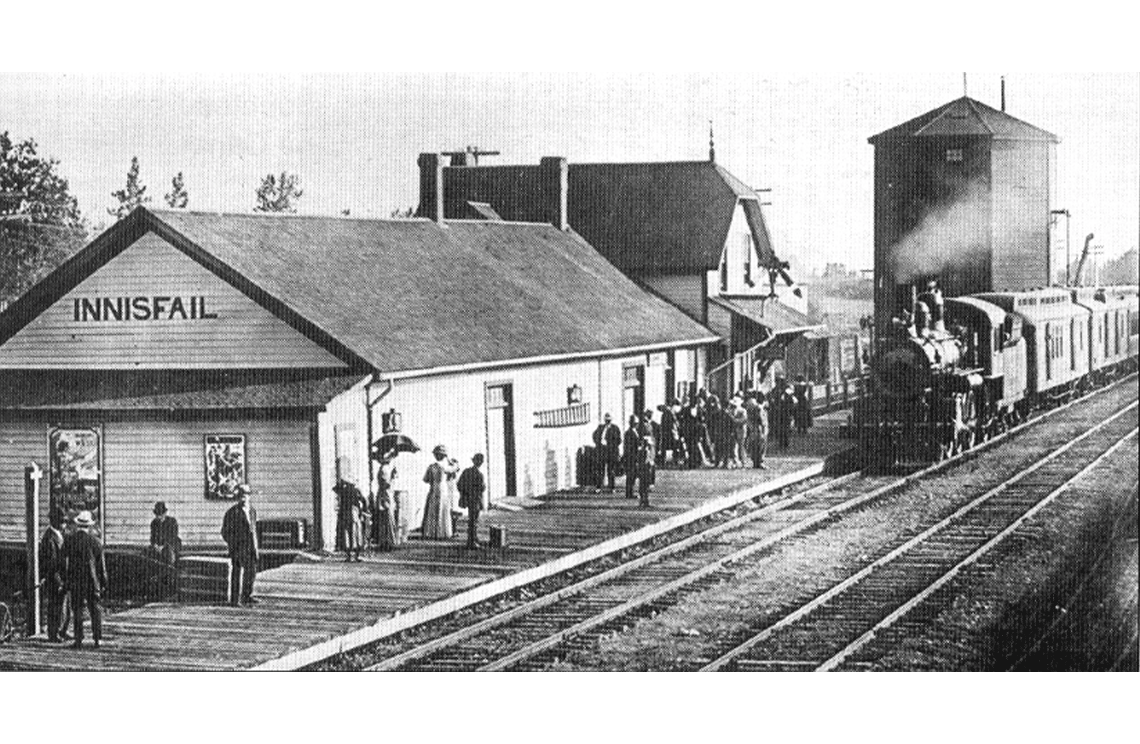 A train pulls into a railway station, people wait on the platform. The station itself has a basic rectangular structure and sloped roof.