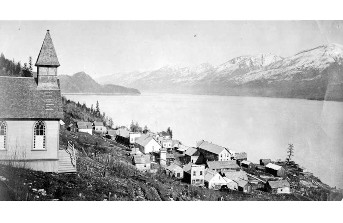 A large church and a cluster of houses and buildings on a hillside next to a lake. Opposite the lake are snow-capped mountains.