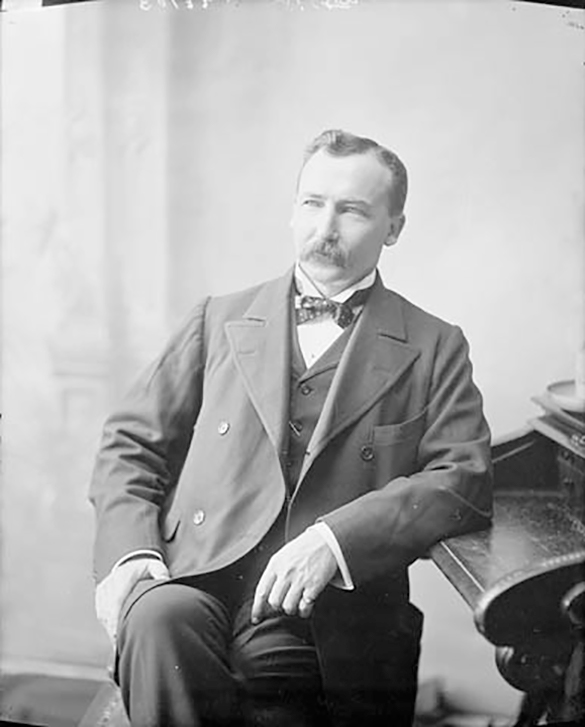 This is Clifford Simon. He is wearing a suit and sitting on a chair leaning on a desk.