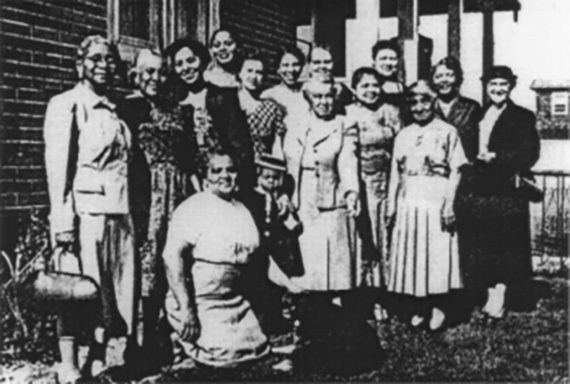 A group of women in dresses smile and pose in front of a brick home.