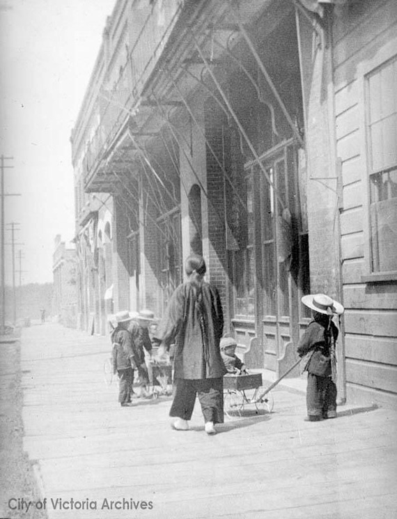 A woman and several children wearing hats with ribbons walking along Chinatown boardwalk