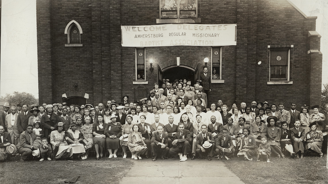 large group of well-dressed people posing in front of a church with a banner reading “Welcome Delegates”