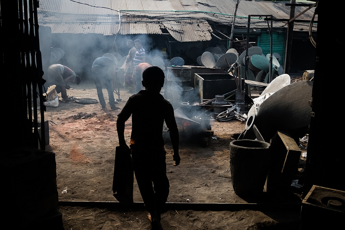 A silhouette of a child walking while carrying something. The ground of the building is sandy.