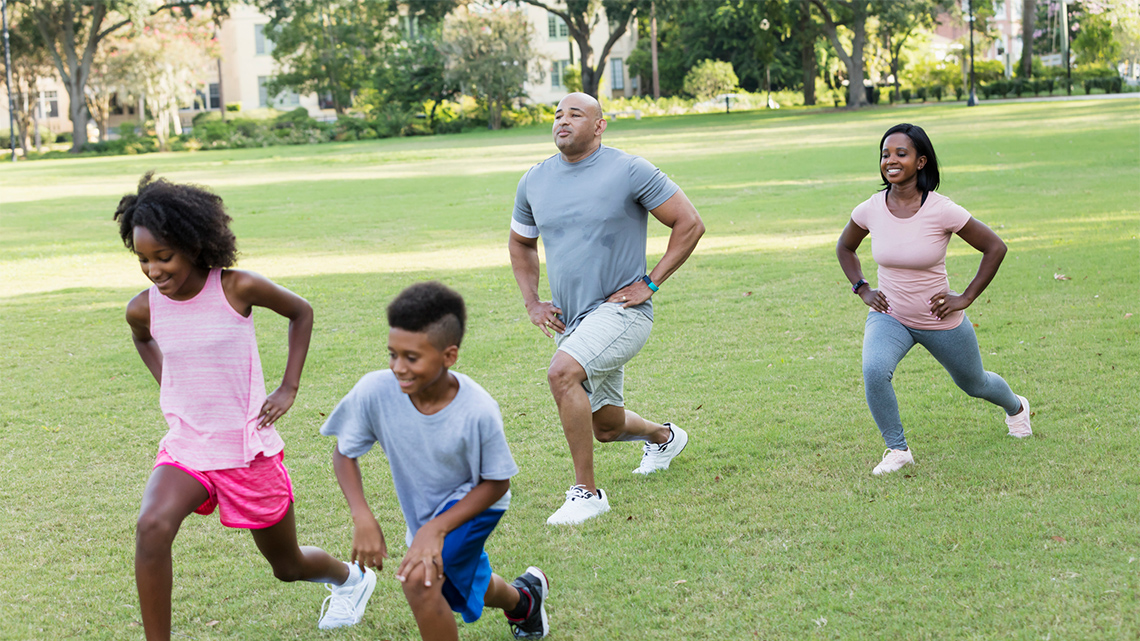 A family of two adults and two children are doing leg lunges on the grass.
