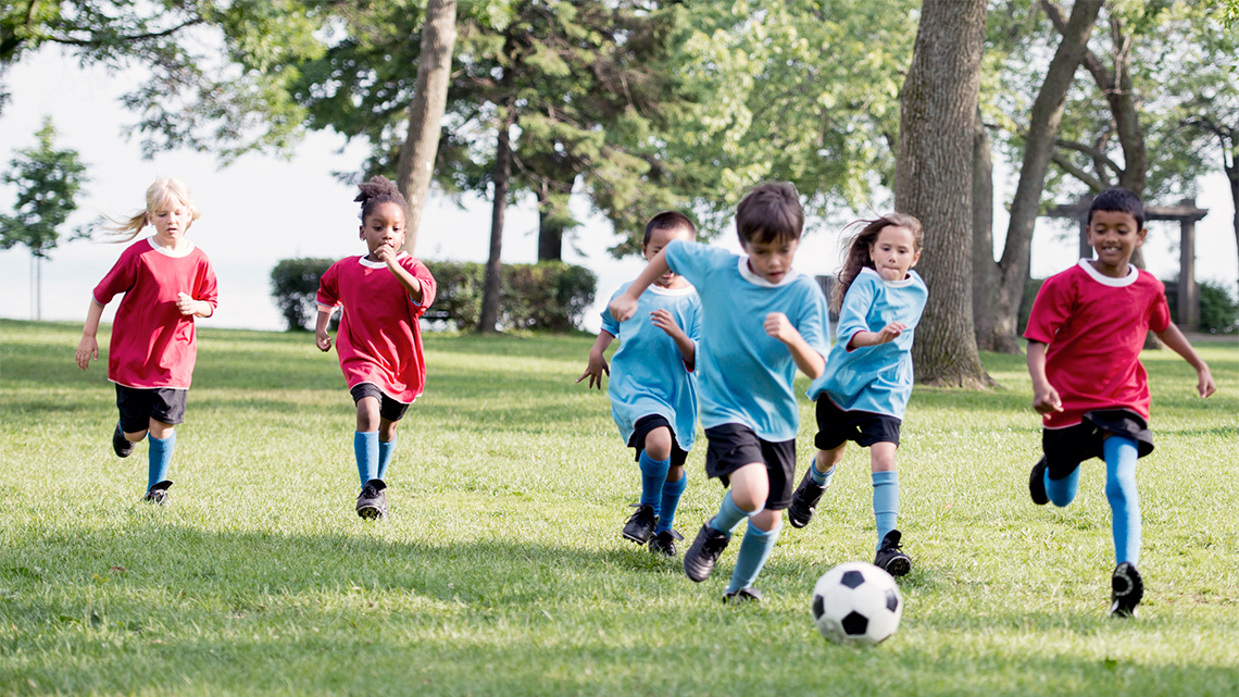 Two teams of children are in soccer uniform, running towards a soccer ball.