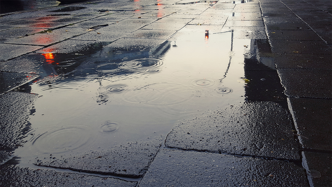 It is raining on a wet sidewalk and it is covered in a large puddle.