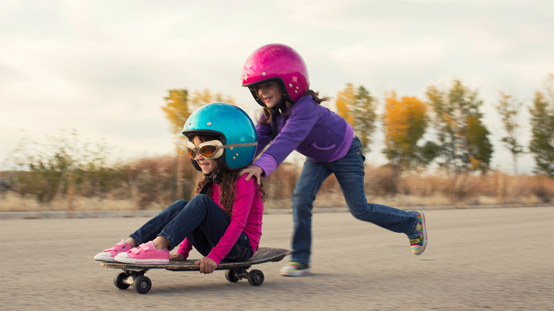 A child is seated on a skateboard and another child is pushing the skateboard. They both wear long sleeve shirts, jeans, running shoes and helmets.