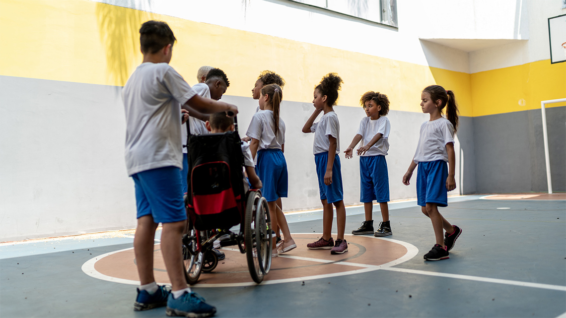 Students in matching t-shirts and shorts are in a gym, standing close together and listening to their teacher.