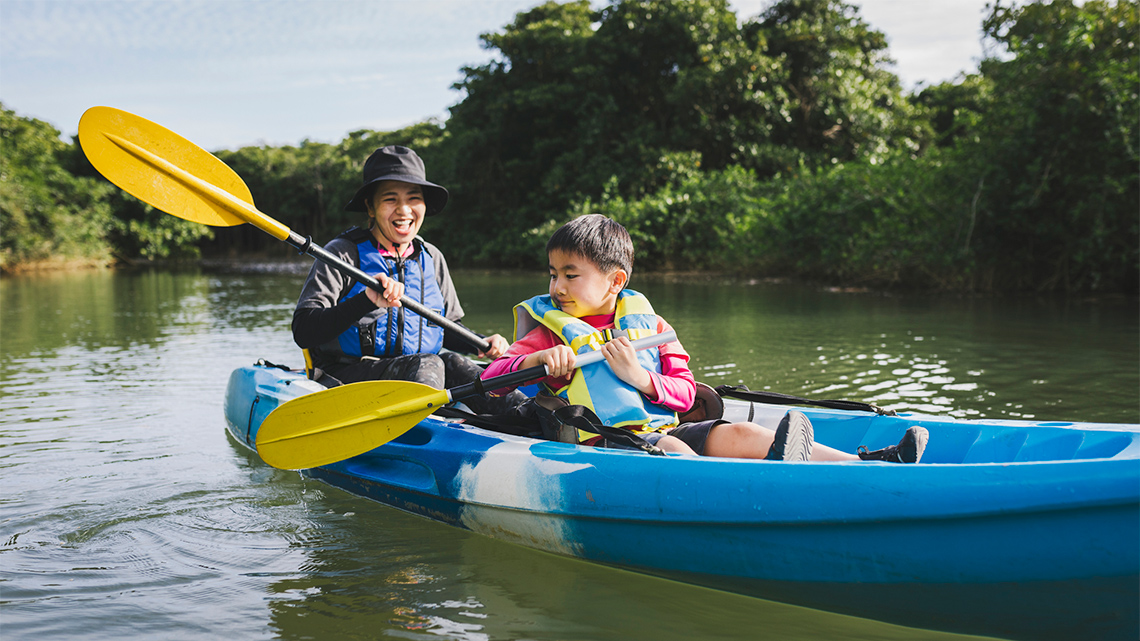 A parent and child in lifejackets are paddling a canoe outdoors.