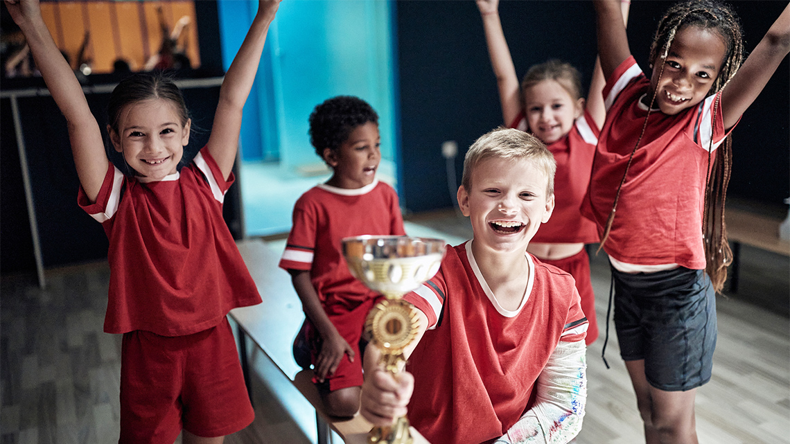 A team of young children have big smiles and raise their hands up while one student on the team holds a trophy. 