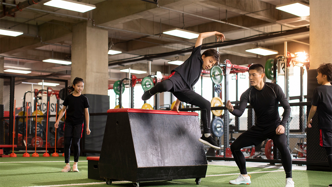 Three children and an instructor are in a gym. One child jumps over a hurdle guided by the adult.