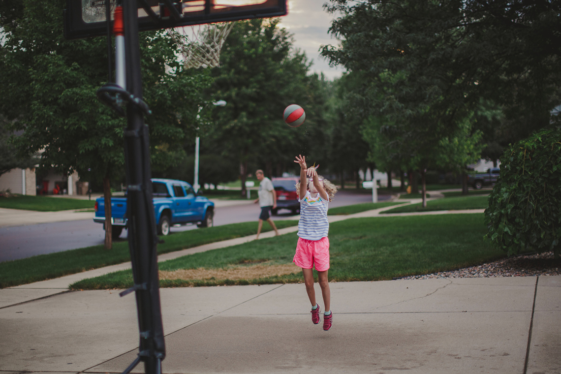 A child is playing basketball in a driveway and trying to get the ball in the outdoor basketball net.