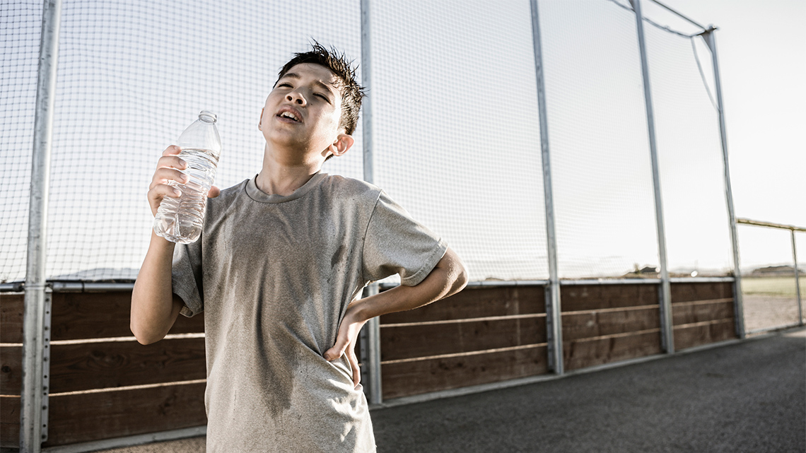 The sun is shining brightly. A child is outside and his t-shirt is wet with sweat. He is holding an uncapped water bottle.