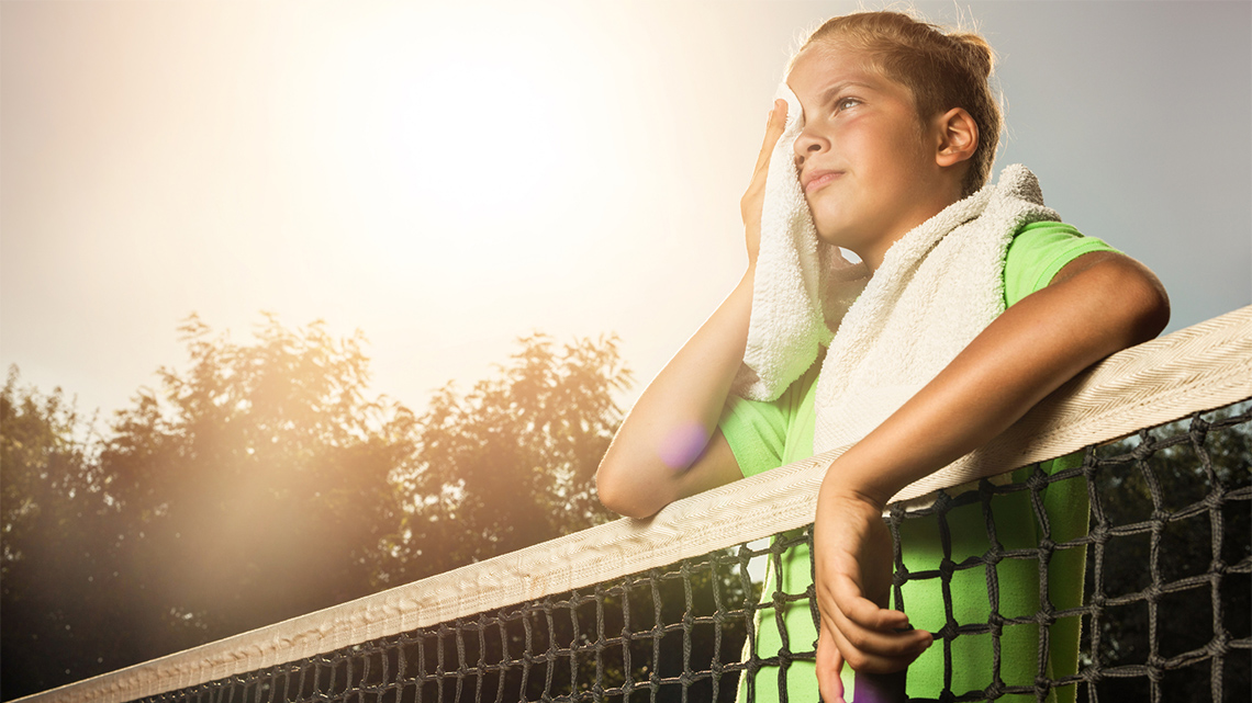 A child stands at a tennis net outside. She uses a towel to wipe sweat from her face.