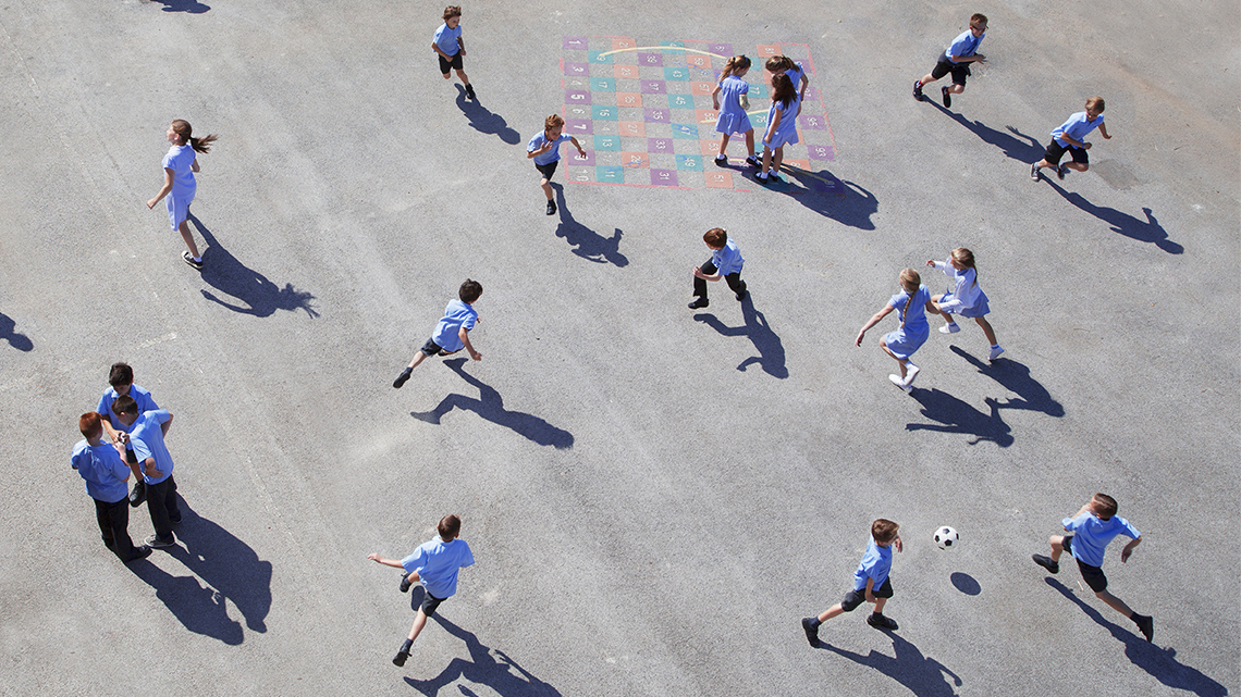 Children playing outdoors in a paved area. Some are playing hopscotch, skipping, running and one is playing with a soccer