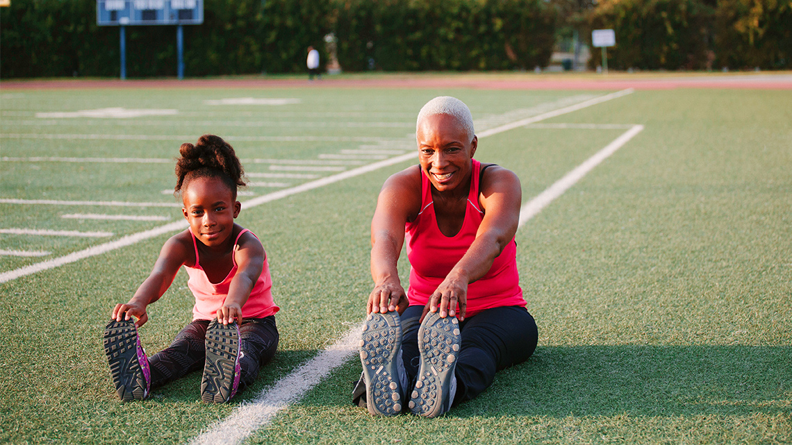 An adult and a child sitting in a sports field with arms touching their toes.
