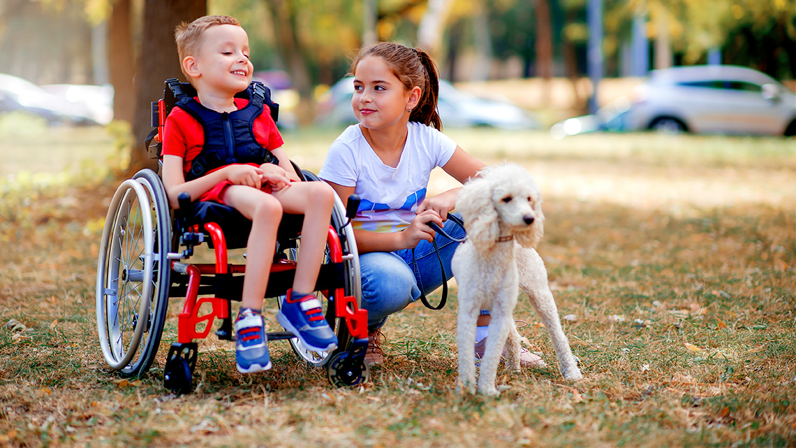 Two children outdoors laughing with a dog. One is in a wheelchair, the other is holding the dog.