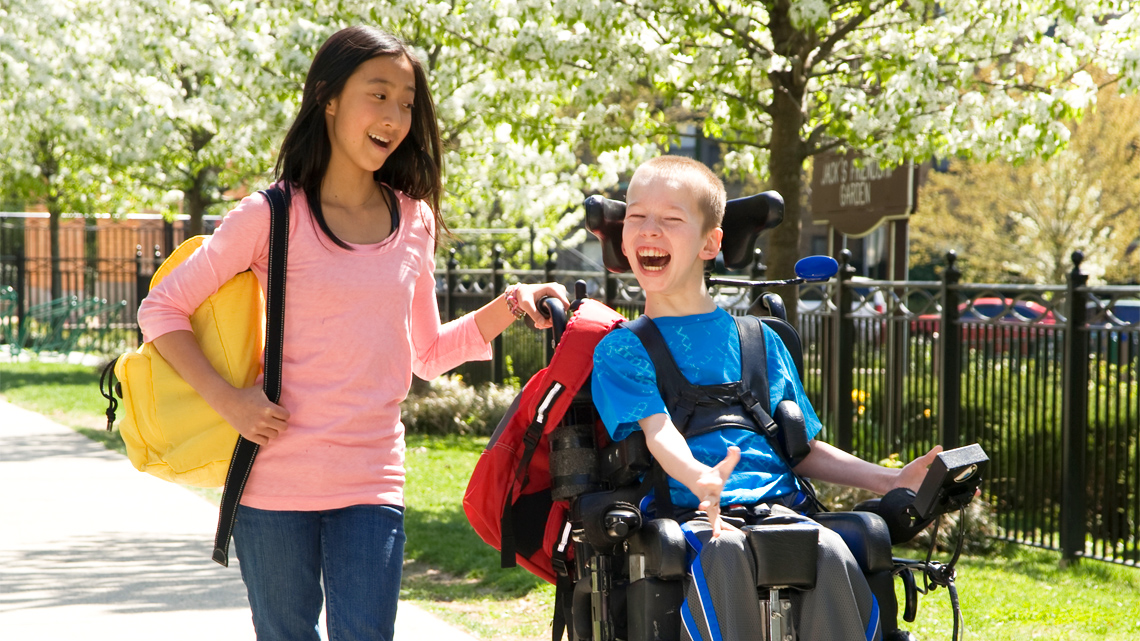 A child in wheelchair and their friend laugh during a walk in a neighbourhood.