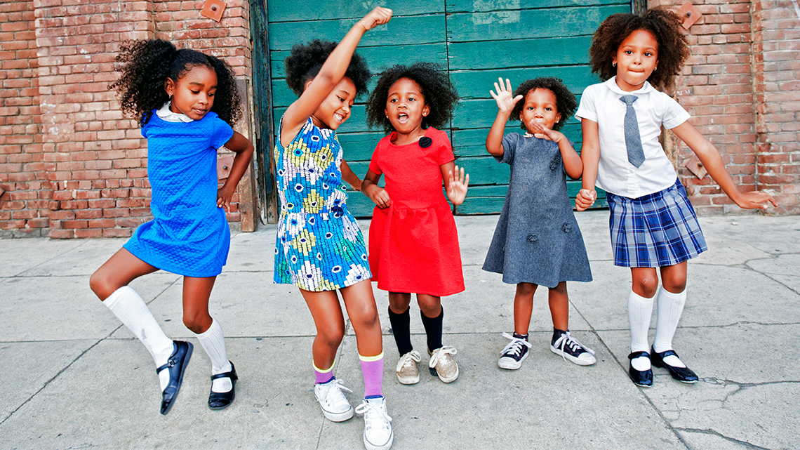 Five children dancing outdoors happily. Two have hands in the air and one has hands on hip.